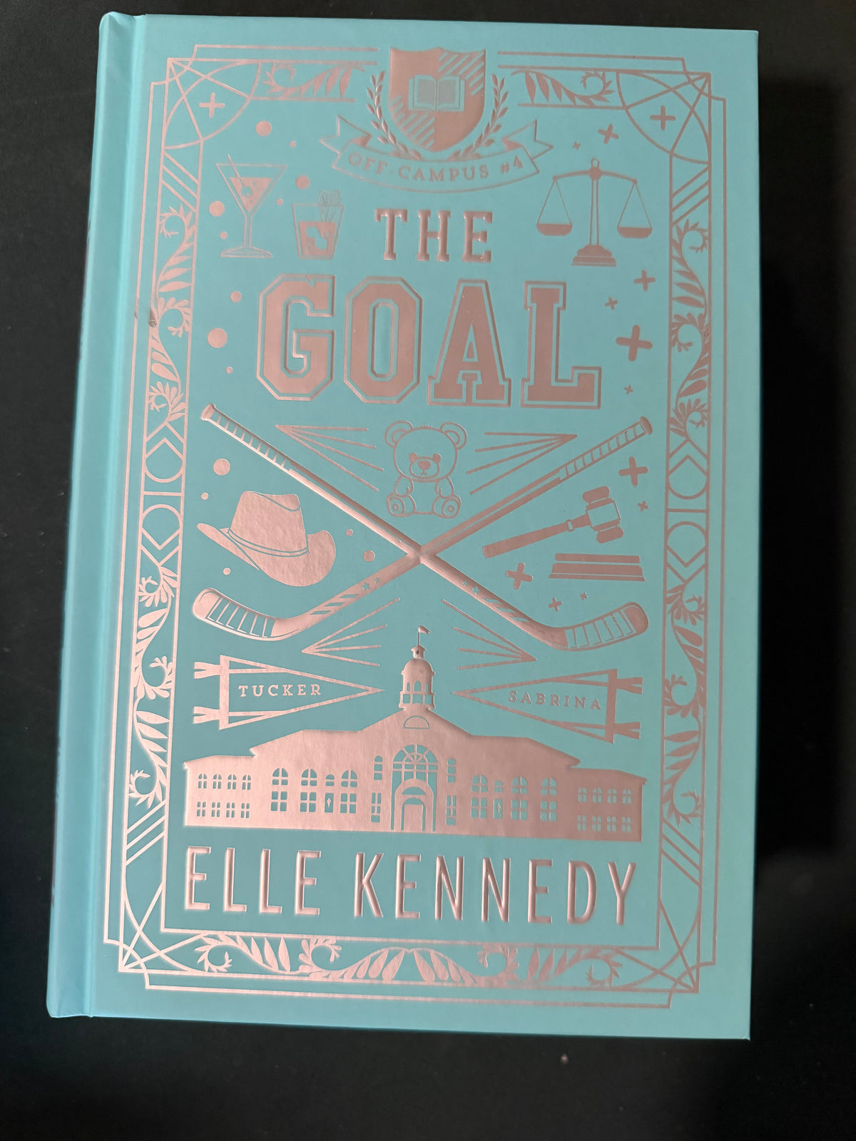The Goal hardcover book by Elle Kennedy, featuring a light blue cover with rose gold foil illustrations and text, photographed on a dark background.
