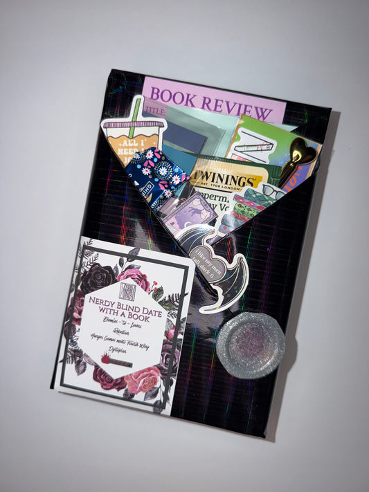 A black mystery book bundle package with a triangular envelope opening filled with themed book review inserts, a 'Nerdy Blind Date with a Book' card, a silver witch hat, and a round glittery seal, photographed on a light gray background.