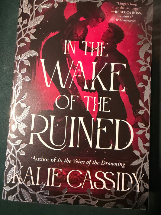 Book cover for 'In the Wake of the Ruined' by Julie Cassidy, featuring a dark red and black background with white gothic typography and a couple embracing in the center.