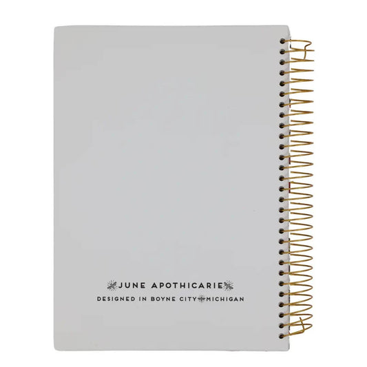 A light gray spiral notebook with a gold wire coil binding and black printed text reading 'JUNE APOTHICARIE' and 'DESIGNED IN BOYNE CITY, MICHIGAN' on the cover, shown on a white background.