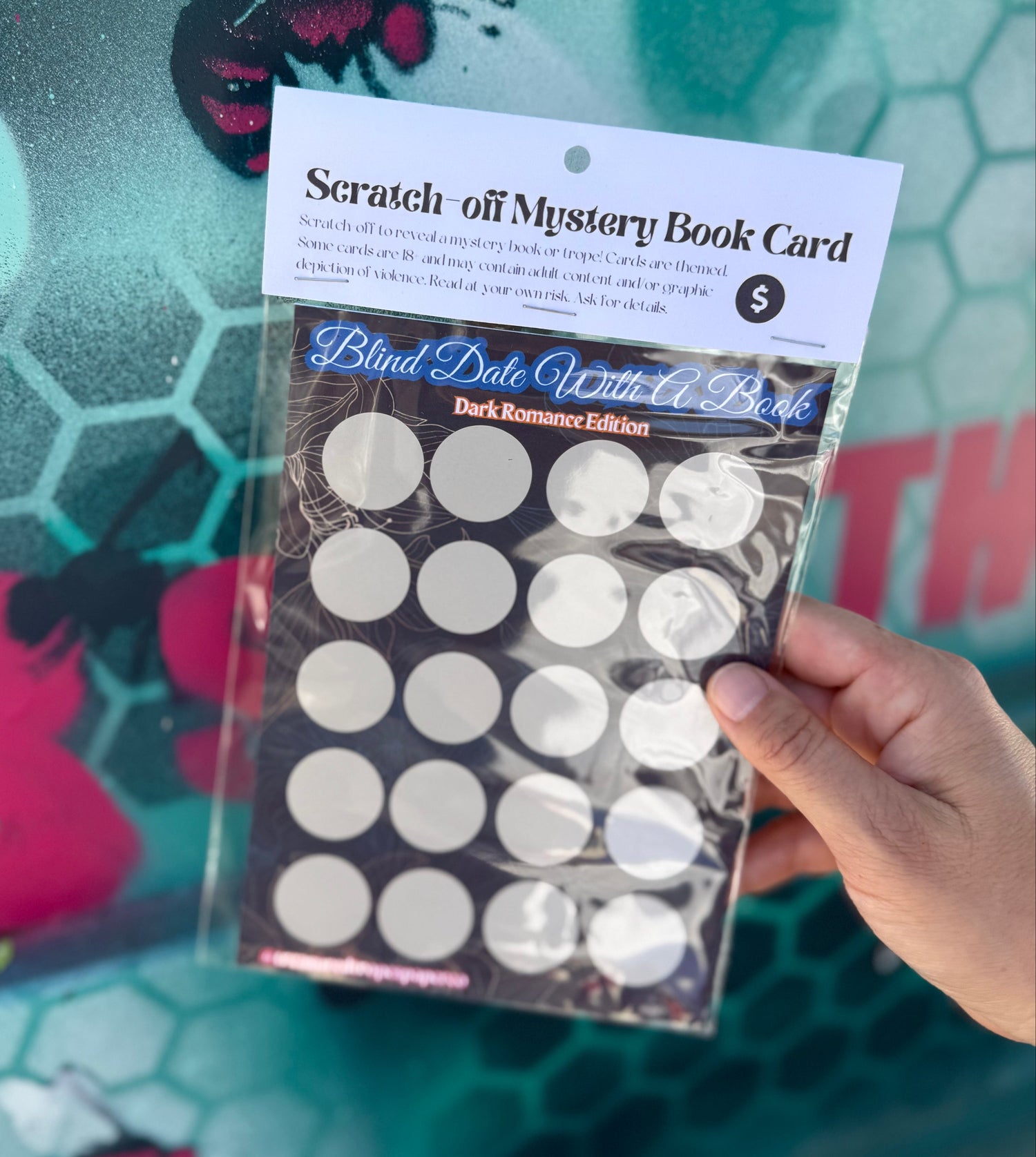 Packaged scratch-off mystery book card held by a hand with a colorful background