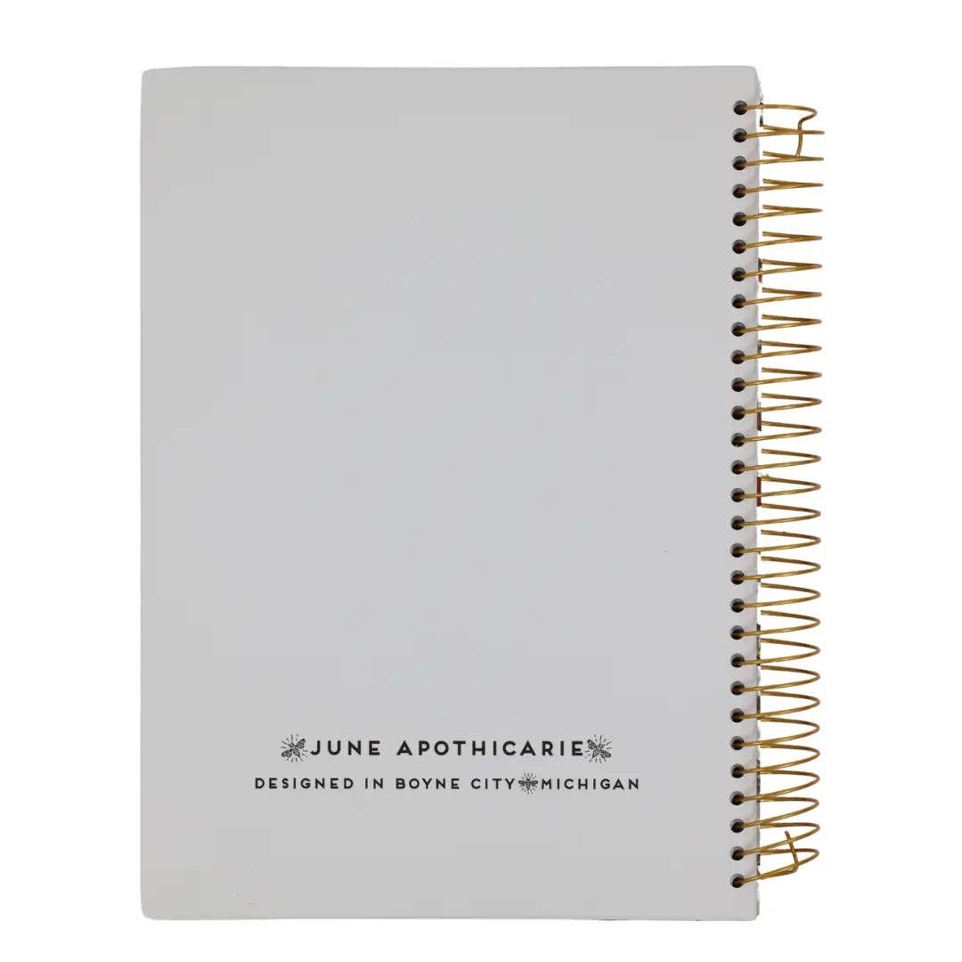 A light gray spiral notebook with a gold wire coil binding and black printed text reading 'JUNE APOTHICARIE' and 'DESIGNED IN BOYNE CITY, MICHIGAN' on the cover, shown on a white background.