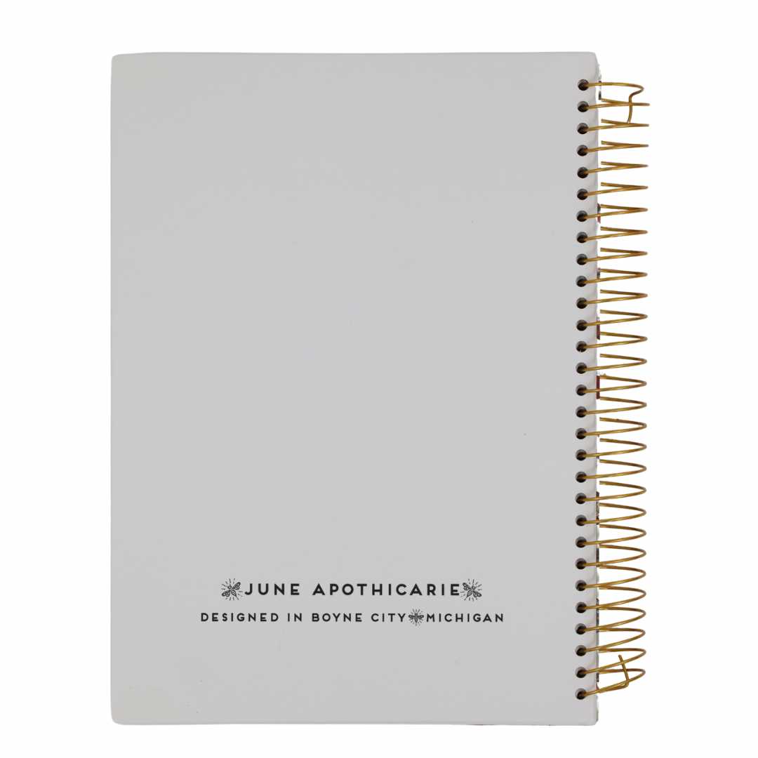 White notebook with gold spiral binding and 'June Apothicarrie' branding on a white background