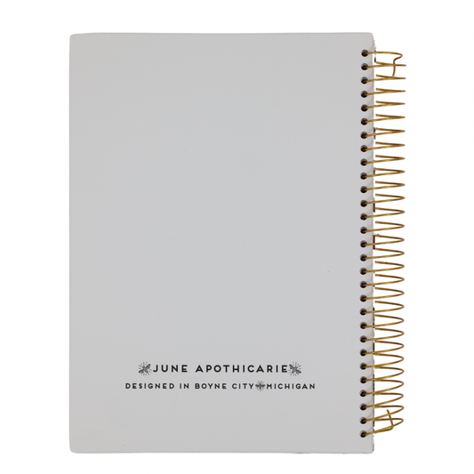 White notebook with gold spiral binding and 'June Apothicarrie' branding on a white background