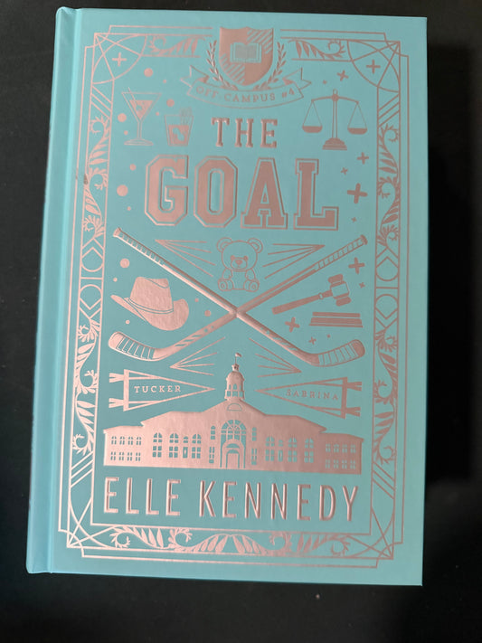 The Goal hardcover book by Elle Kennedy, featuring a light blue cover with rose gold foil illustrations and text, photographed on a dark background.