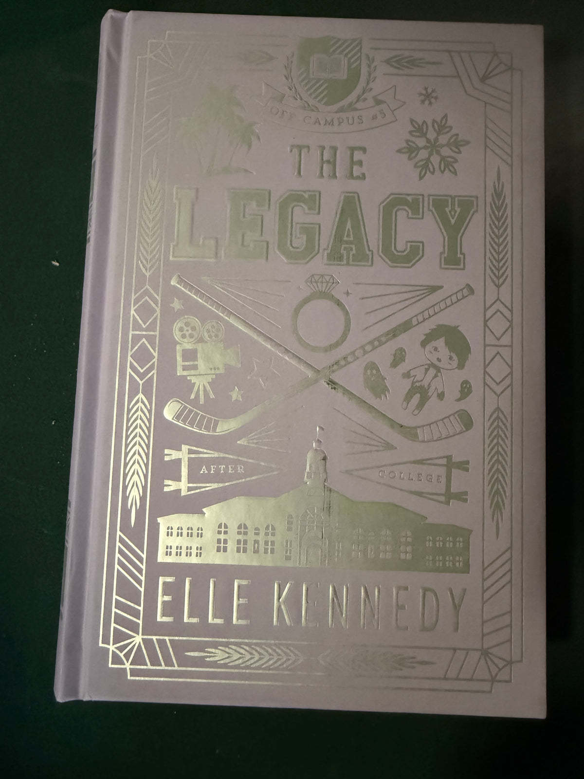 The Legacy hardcover book by Elle Kennedy, featuring a pale lavender cover with metallic gold foil illustrations and text, photographed on a dark green background.